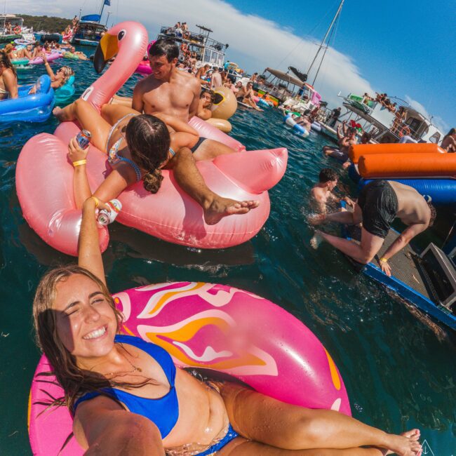 A woman in a blue bikini smiles for a selfie on a pink float in crowded, sunny waters with people on colorful inflatables and boats in the background.