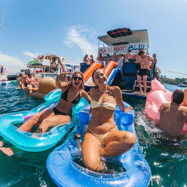Two women in swimsuits smile and pose on blue inflatable loungers in the water at a lively boat party, with people socializing and relaxing on floats and a houseboat in the background under a sunny sky.