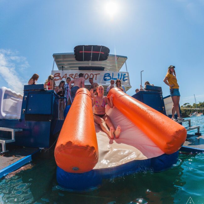 A woman slides down an inflatable orange and white water slide attached to a boat named "Barefoot Blue," surrounded by people enjoying a sunny day on the water.