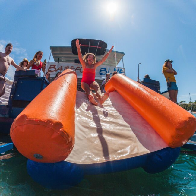 A woman in a red swimsuit slides down an inflatable water slide from a boat into the water, smiling with arms raised. People watch and take photos under a bright sunny sky.