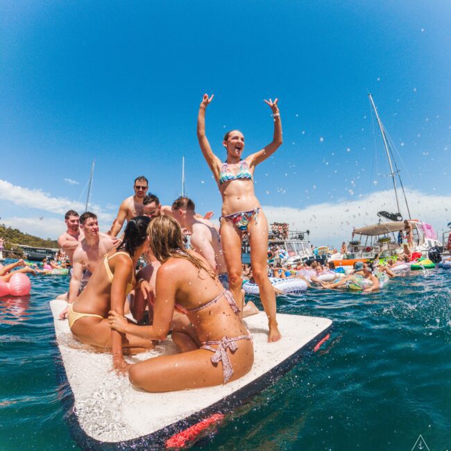 A group of people in swimsuits gather on a floating mat in the ocean, smiling and playing in the sun. One woman stands with arms raised, surrounded by boats and others enjoying the water.