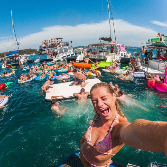 A woman in a bikini takes a smiling selfie on a boat, surrounded by people on floats and boats, enjoying a lively party on bright blue water under a sunny sky.