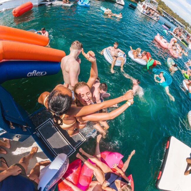A group of people enjoys a lively pool party on a lake, swimming and floating on colorful inflatables near boats. Two women in swimsuits laugh and hug at the edge of a boat as others relax in the water.