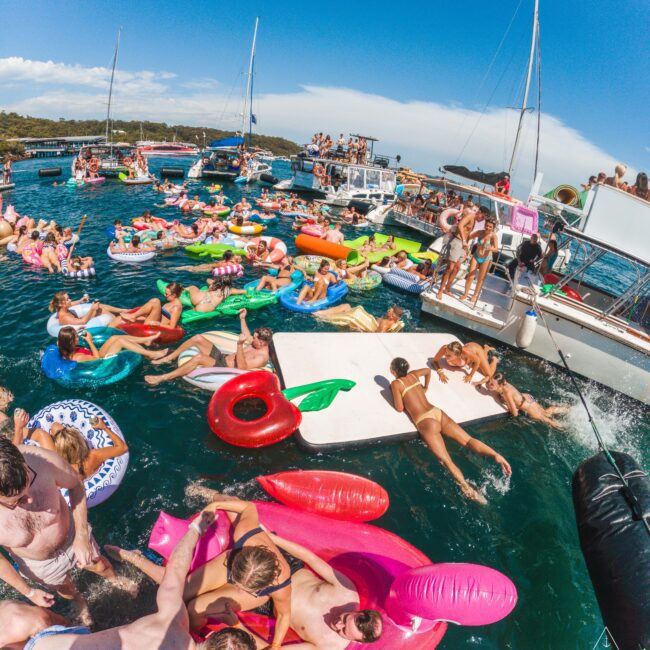 A lively boat party with many people swimming, relaxing on colorful pool floats, and gathering on yachts under a bright blue sky. The scene is vibrant and energetic, with lots of activity in and around the water.