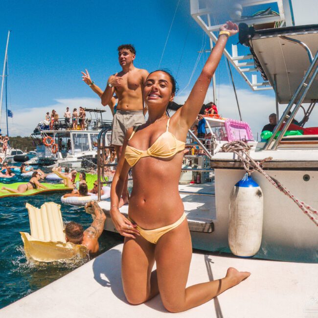 A woman in a yellow bikini poses and smiles on a boat, raising one arm. Behind her, people enjoy a lively party on multiple boats, with floats in the water under a sunny sky.