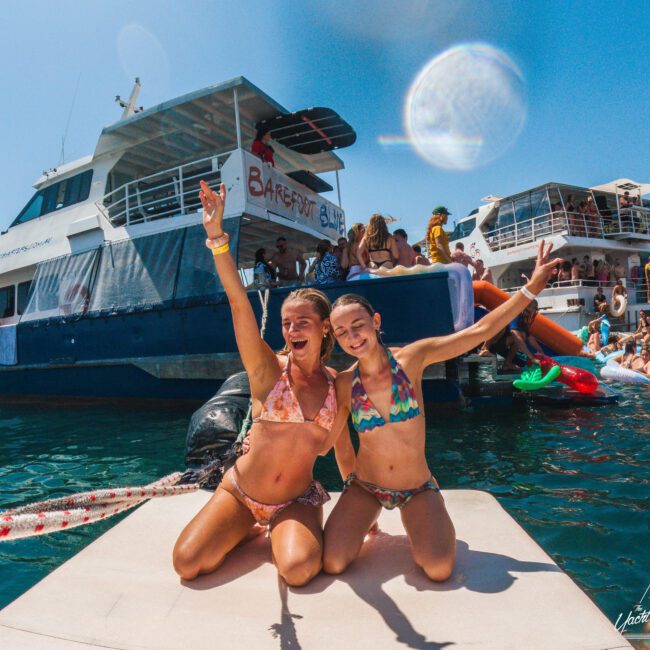 Two smiling women in bikinis kneel on a dock, posing with raised arms in front of boats and a lively crowd enjoying a sunny day on the water. Inflatable toys and people swimming are visible in the background.