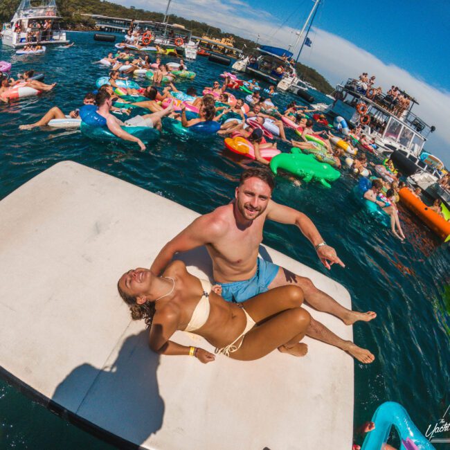 A man and woman relax and laugh on a floating platform in the water, surrounded by many people on colorful inflatables, with boats and a lively crowd in the background under a sunny sky.