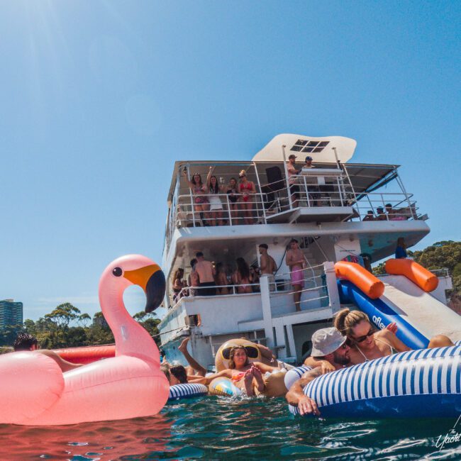 People relaxing on colorful pool floats, including a large pink flamingo, near a white party boat with others onboard. It’s a sunny day and everyone appears to be enjoying the water and festivities.