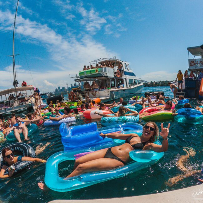 People relax on colorful pool floats in a crowded, lively scene on the water near boats under a bright blue sky. The atmosphere is festive, with many swimmers and sunbathers enjoying a sunny day.