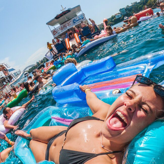 A woman in a bikini smiles and sticks out her tongue while lounging on a colorful float in a crowded pool party; many people are floating nearby, with a party boat and city buildings in the background.