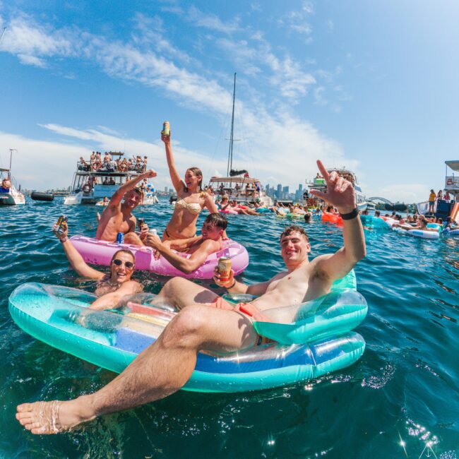Five people relax on colorful inflatable rafts in the water, smiling and holding drinks, with boats and more people celebrating in the background under a sunny blue sky.