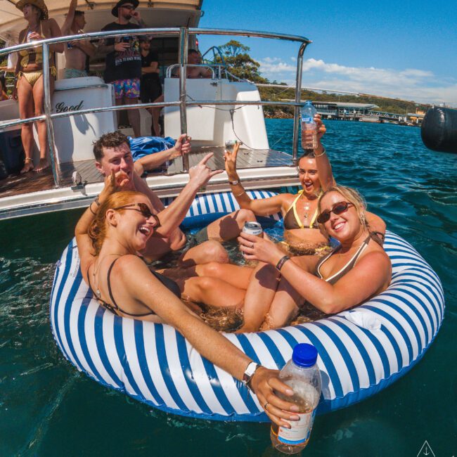Four people in swimsuits relax and smile on a striped inflatable raft in the water, holding drinks and posing for the camera. A boat and others are visible in the background on a sunny day.