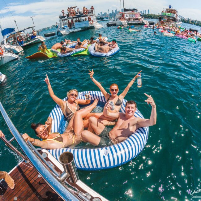 Four people in swimsuits relax and smile on a striped inflatable float, holding drinks, surrounded by boats and others swimming and lounging in a lively, sunny floating party scene on blue water.