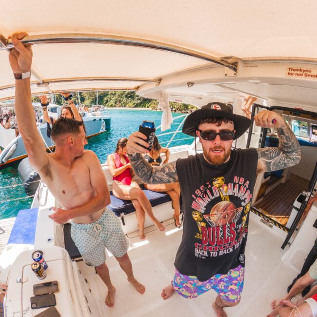 A group of people enjoying a sunny day on a boat; a man in sunglasses and a Chicago Bulls shirt poses and flexes his arms for a selfie, while others relax and chat around him.
