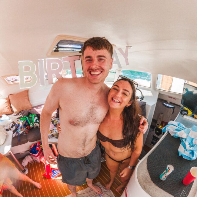 A smiling man and woman in swimsuits stand close together on a boat decorated with "BIRTHDAY" letters. The boat interior appears festive, with drinks and swim gear scattered around.