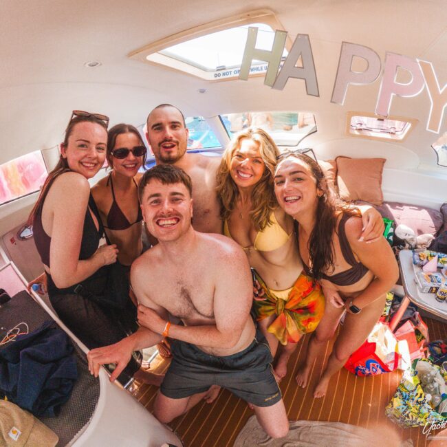 A group of six young adults in swimsuits smile and pose together inside a boat. Festive "HAPPY" letters hang above them, and sunlight shines through the windows, giving a cheerful, vacation vibe.