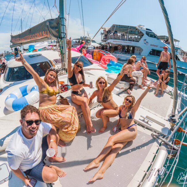 A group of people in swimsuits and sunglasses smile and pose on a boat during a sunny day party. Other boats and people are visible in the background on the water, with colorful pool floats and a festive atmosphere.