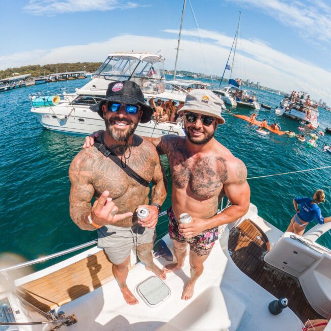 Two tattooed men in swim shorts and hats stand smiling on a boat, holding drinks. Behind them, people relax on other boats and inflatables on the water under a blue sky. It appears to be a lively summer gathering.
