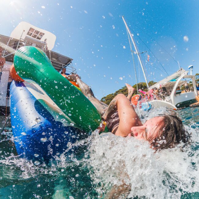 A person splashes into the water from an inflatable float near boats on a sunny day, with droplets frozen midair and blue sky in the background.