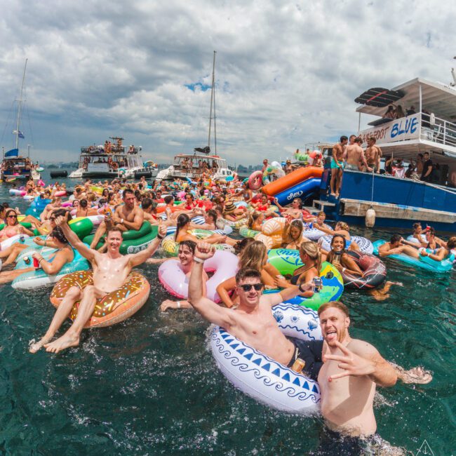 A large group of people in colorful inflatable tubes and floats enjoy a lively party in the water near several boats under a partly cloudy sky. Many are smiling, posing, and having fun in the festive atmosphere.
