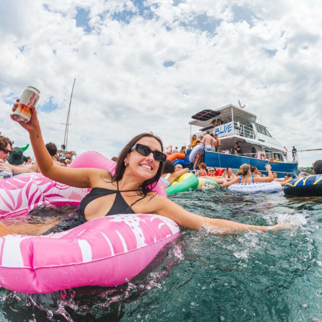 A woman in sunglasses smiles while floating on a pink inflatable in the water, holding a drink. Around her, people on inflatables gather near a boat under a cloudy sky, enjoying a lively outdoor party.