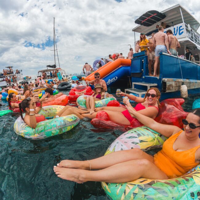 People relax on colorful inflatable floats in the ocean near a docked boat. Some are smiling, taking photos, and enjoying the sunny day, surrounded by other boats and a lively, festive atmosphere.
