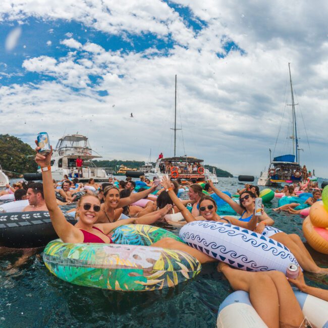 A group of young adults relax on colorful inflatables, smiling and raising drinks while floating on the water near boats under a partly cloudy sky. The atmosphere is lively and festive.