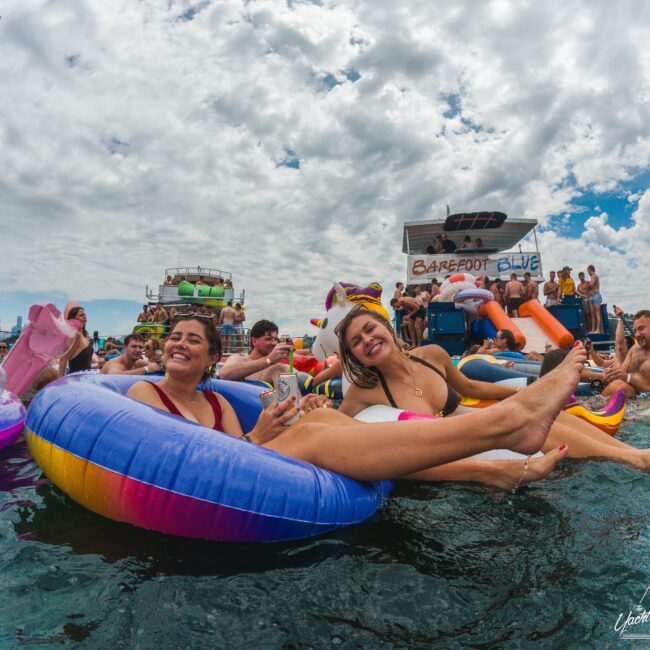 Two women in colorful floaties smile and relax in the water during a crowded outdoor party, surrounded by other people on inflatables with boats and a "Barefoot Blue" sign in the background under a cloudy sky.
