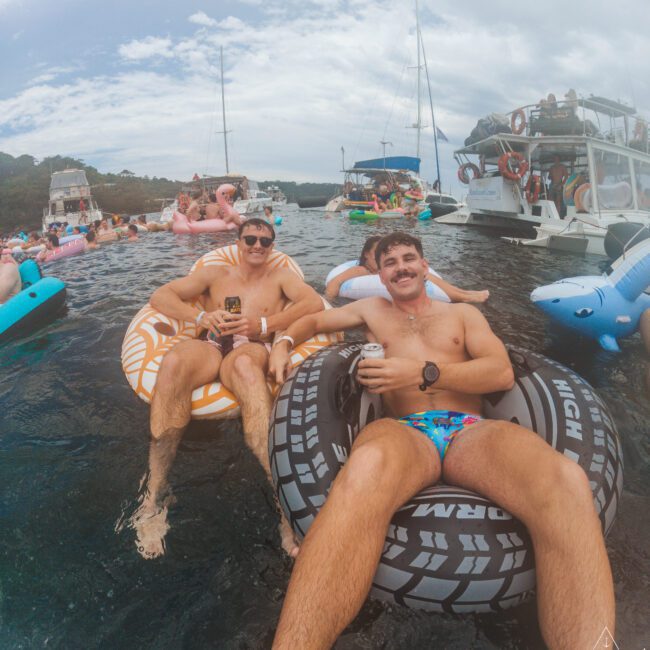 Two smiling men in swim trunks relax on inflatable tubes, holding drinks in a crowded bay. Other people float nearby, surrounded by boats and inflatable pool toys under a cloudy sky.