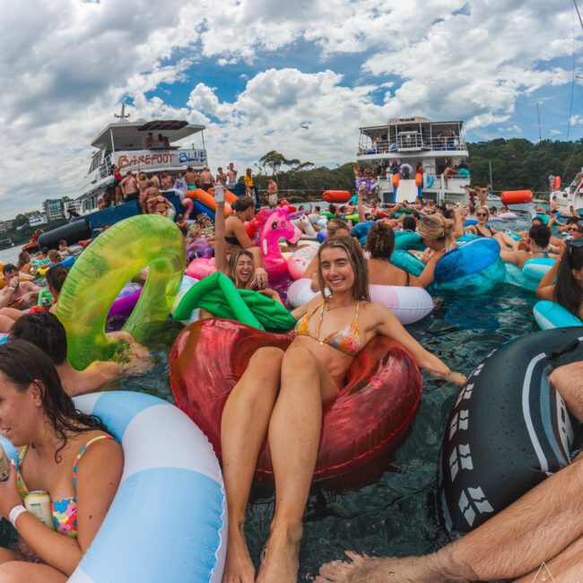 A woman in a bikini relaxes on a red pool float among a crowd of people on colorful inflatables in the water, with boats and a cloudy sky in the background.