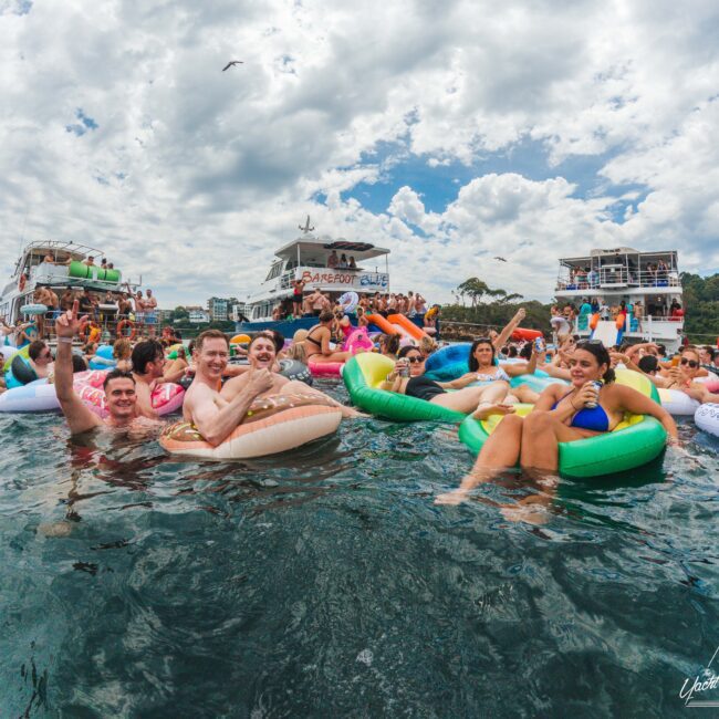 A group of people relax on colorful inflatable tubes in the water near boats during a lively party under a partly cloudy sky, with others seen in the background enjoying the festive atmosphere.