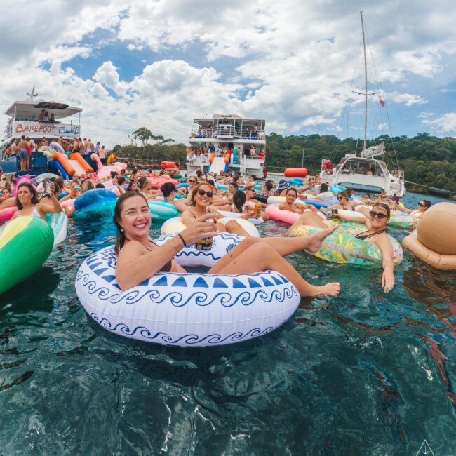 A group of people relax on colorful inflatable floats in the water near boats, smiling and enjoying a sunny day with partly cloudy skies. The atmosphere is lively and festive.