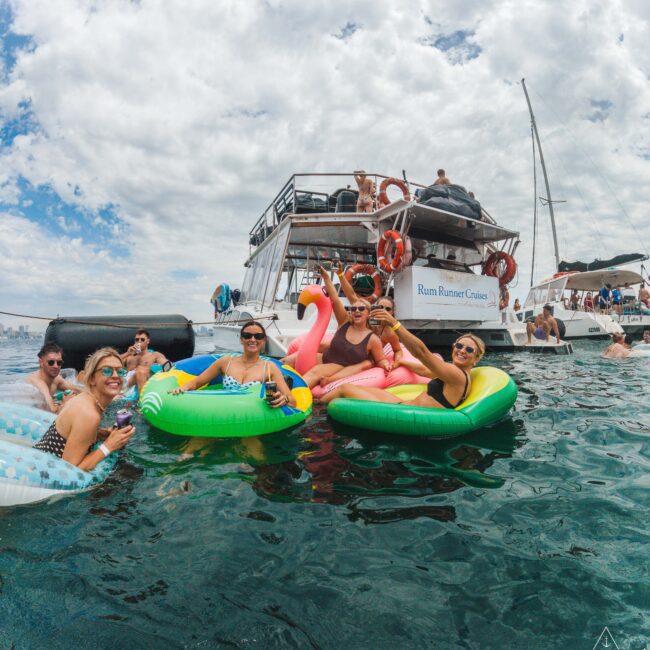 A group of people relax on colorful pool floats in the water near a large boat, enjoying a sunny day. The sky is partly cloudy, and others are visible on the boat and in the water nearby.