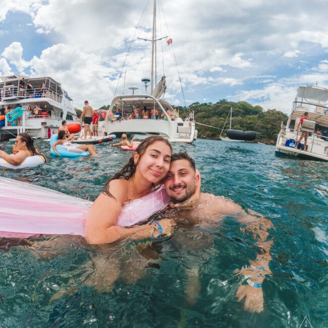 A couple smiles while relaxing on a pink float in the ocean, surrounded by other people swimming and lounging near anchored yachts on a partly cloudy day.