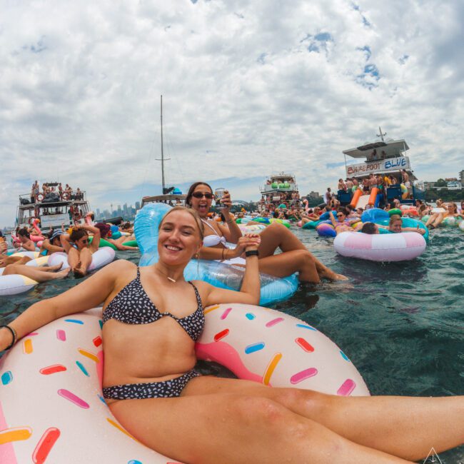 A woman in a polka dot bikini smiles while lounging on a donut-shaped float in the water, surrounded by other people on colorful inflatables at a lively boat party under a cloudy sky.