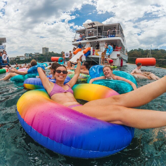 People relaxing on colorful inflatable tubes in the water near a docked yacht, with others enjoying the scene under a partly cloudy sky; a festive, social summer atmosphere.