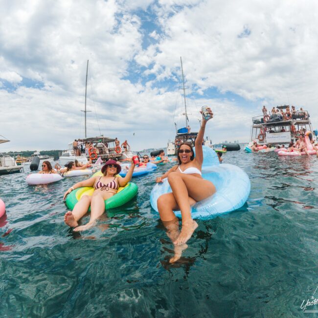 People relax on colorful inflatable floats in the water near several boats. A smiling woman in sunglasses raises a can, surrounded by others enjoying the sunny, festive atmosphere. The sky is partly cloudy.
