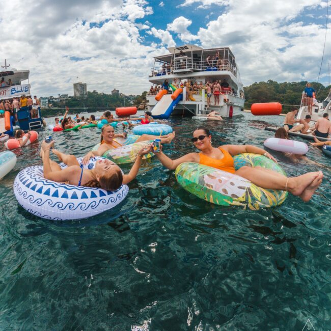 Two women in colorful inflatable tubes relax on the water, clinking drinks, surrounded by others enjoying a lively party near a large boat under a partly cloudy sky.