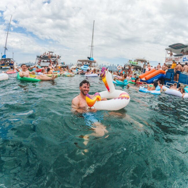 A man smiles while floating on a unicorn pool float in the ocean, surrounded by people on colorful inflatables and boats under a partly cloudy sky. A city skyline is visible in the distance.