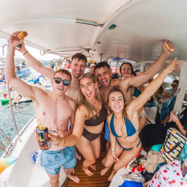 A group of six smiling young adults in swimwear pose with their arms raised on a boat, surrounded by snacks and drinks, with water and other boats full of people visible in the background.