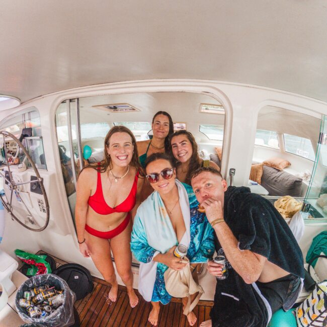 Five people in swimwear and towels stand smiling together on a boat, appearing happy and relaxed. The boat's interior, some seats, and scattered cans are visible in the background.