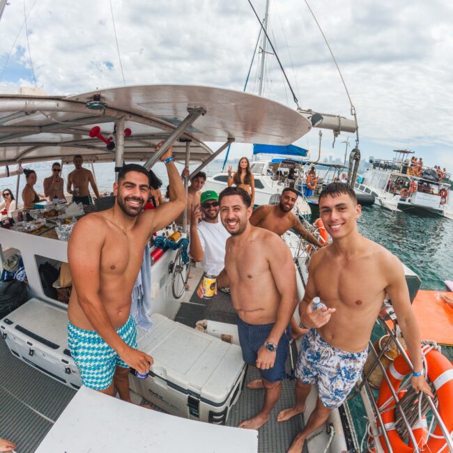 A group of smiling young men in swimwear stand together on a boat, posing for the camera. Other people are enjoying themselves in the background, with the sea and other boats visible under a cloudy sky.