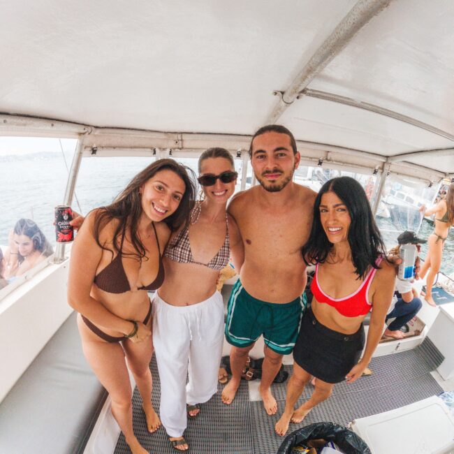 Four young adults in swimwear stand and smile on a boat deck, with the ocean visible in the background. The group appears relaxed and is enjoying a sunny day on the water.