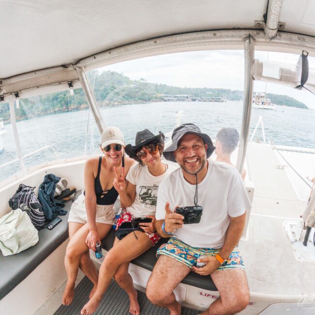Three smiling people in casual summer clothes and hats sit on a boat, with water and a forested coast visible in the background. One person holds a camera, and another flashes a peace sign.