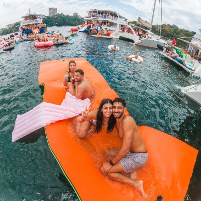 A group of young adults sits and smiles on an orange floating mat in the water, surrounded by boats and people enjoying a lively outdoor party. The sky is partly cloudy and the atmosphere is festive.