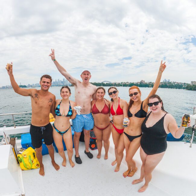 Six smiling people in swimsuits stand on a boat, raising their arms and drinks in celebration. The water and a city skyline are visible in the background under a partly cloudy sky.