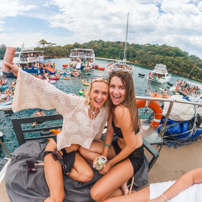 Two young women sit on a boat, smiling and holding drinks, with a lively crowd swimming and relaxing on nearby boats in the background. The scene is festive, with clear water and green trees visible.