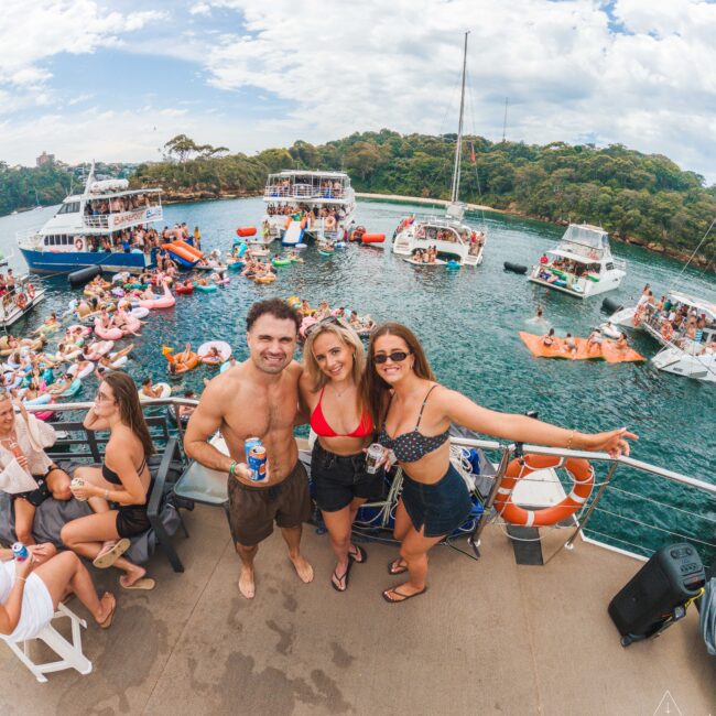 Three friends in swimsuits pose and smile on a boat with drinks in hand, surrounded by other boats and people on inflatables enjoying a lively party on the water under a partly cloudy sky.
