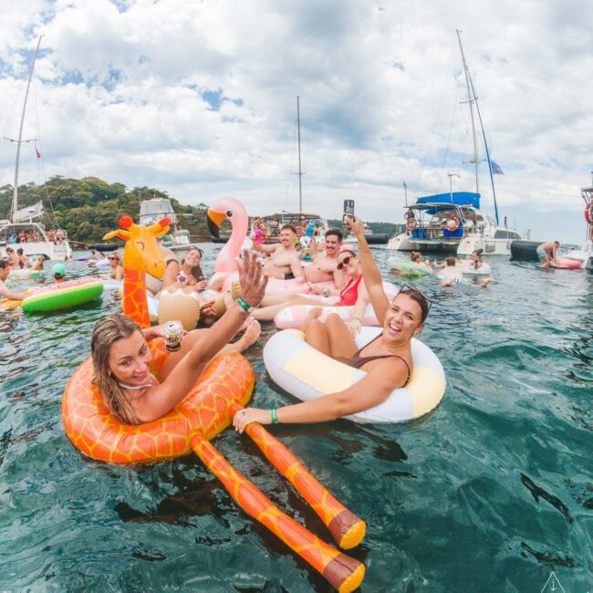 A group of people enjoying a party on colorful inflatable floats in the water, raising drinks and smiling, with boats and a cloudy sky in the background.