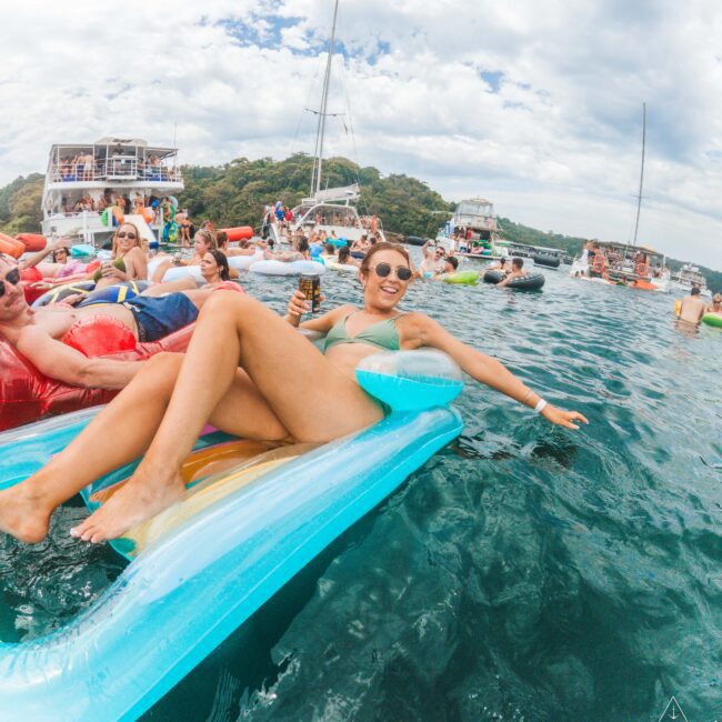 A smiling woman in a green bikini relaxes on a colorful float in clear blue water, surrounded by boats and people on inflatables, enjoying a sunny day at a lively beach party.